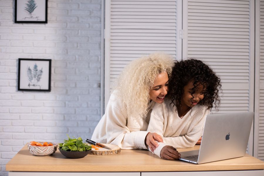 A smiling mother and her daughter look something up together on a laptop at the kitchen counter